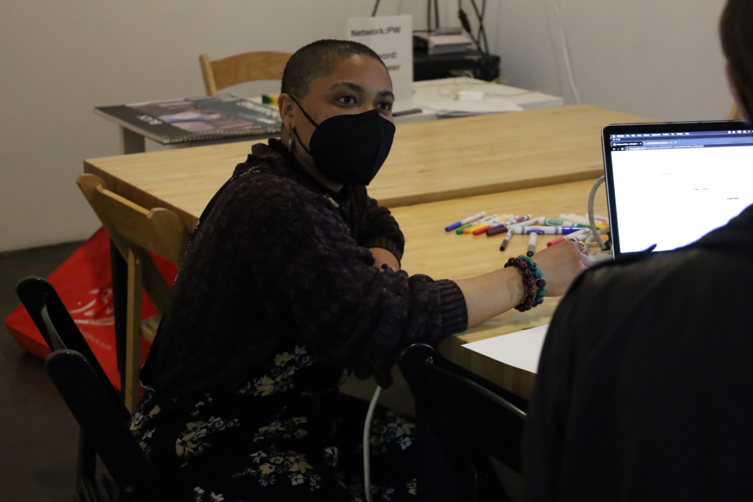 Aiyo O’Connor, who has a shaved head and wears a black mask, a thick sweater and a loose overall with floral patterns, is engaging with a workshop participant off camera. There are a bunch of colored markers on the table.