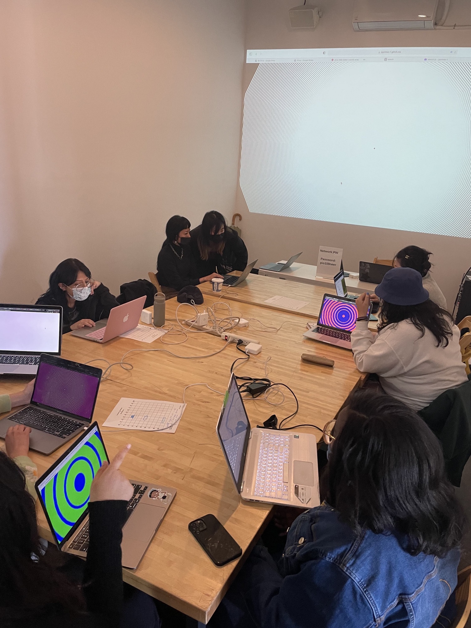 Workshop participants work on their laptops around a long rectangular table. Their laptop screens show remixes of the circular visual pattern projected on the wall.