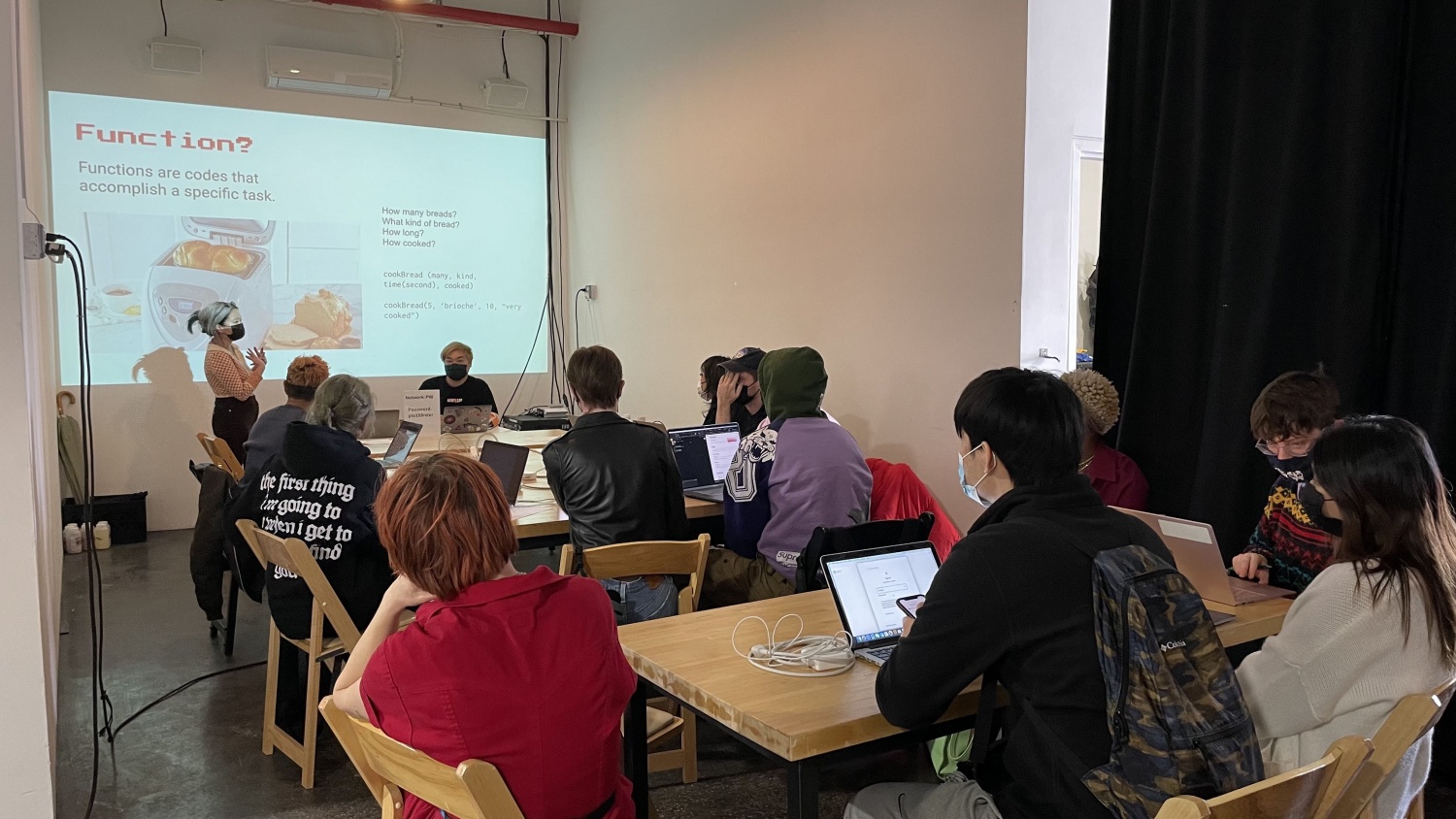 A group of 11 workshop participants sit around a long rectangular table while listening to Iley Cao and Munus Shih going over “function”, a computer programming concept. The projection on the back wall reads “Functions are codes that accomplish a specific task. How many breads? What kind of bread? How long? How cooked?”, accompanied by an image of a bread making machine.