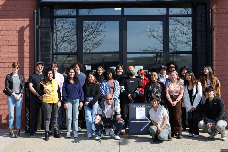 A group of 22 people are in front of a brick building with large glass doors. They stand around a storefront sign that reads &ldquo;Pioneer Works. Code, Decolonized with Power Plant&rdquo; and smiled into the camera.