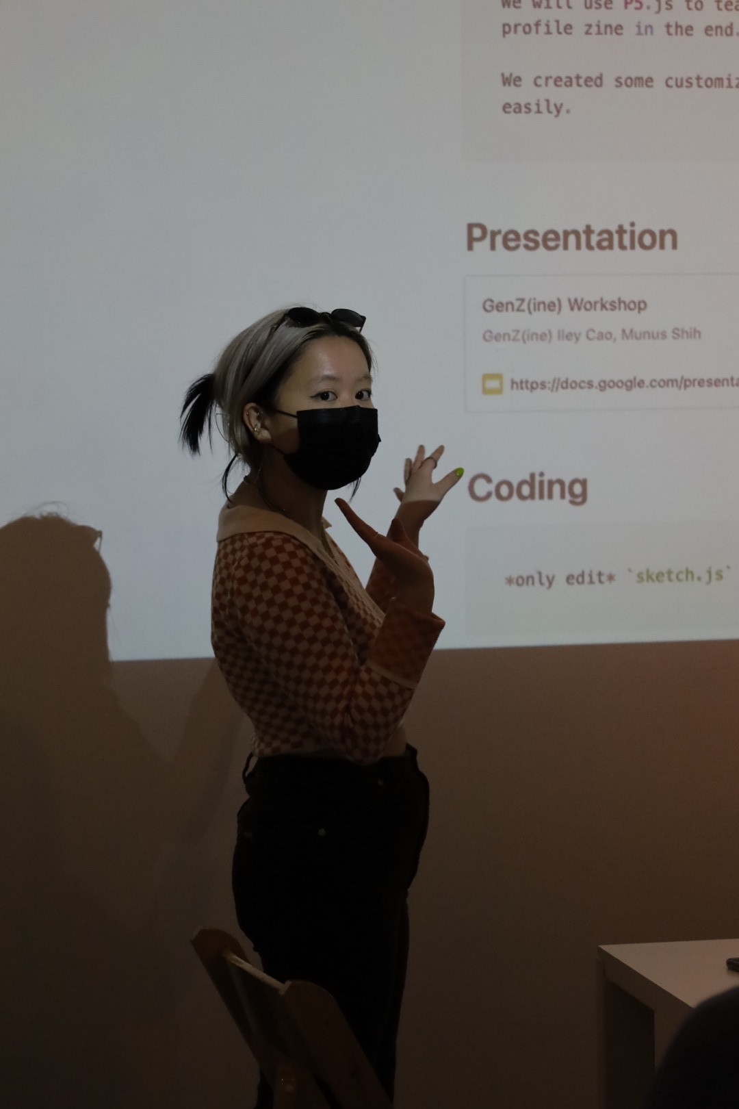 Iley Cao, who has bleached straight hair tied to a low ponytail, is wearing a black mask, a sweater with red and white checkerboard pattern, and sunglasses on her head. She is lighted by the projection of a workshop she is co-leading.