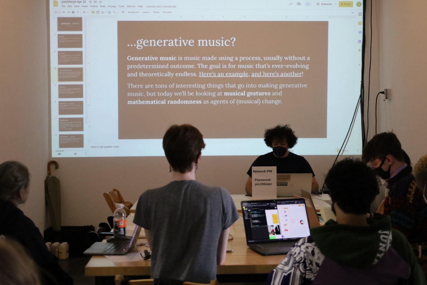 Jay Tobin, who has curly hair and wears a black mask and t-shirt, is teaching a workshop in front of a group of participants. Behind him the presentation slides reads “generative music is music made using a process, usually without a predetermined outcome. The goal is for music that’s ever-evolving and theoretically endless. Here’s an example, and here’s another! There are tons of interesting things that go into making generative music, but today we’ll be looking at musical gestures and mathematical randomness as agents of (musical) change.”