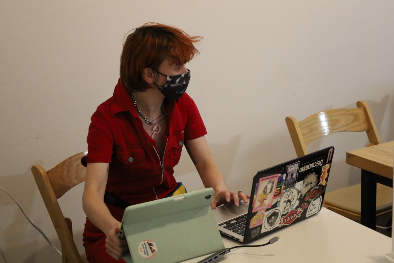 tee topor, who has short orange hair and bangs, wears a black and white tie-dye mask, red jumpsuit and a necklace adorned by a pink hammer and sickle pendant. They are sitting in front of a laptop covered by stickers and a tablet with a light green cover.