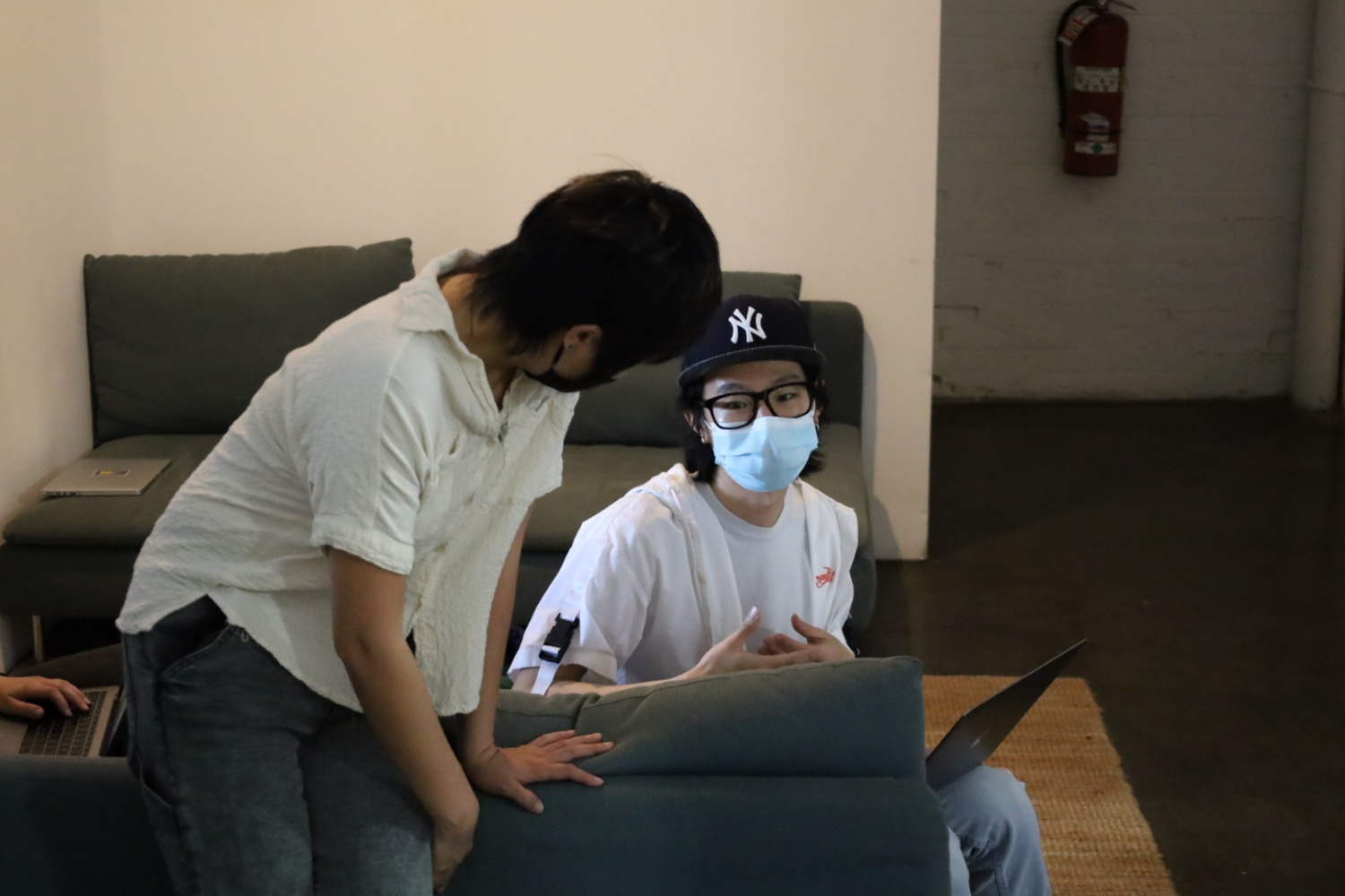 Xin Xin, who has short straight hair and wears a off-white blouse, is leaning against a green couch while talking to Roy Yang, who wears a mask and a New York baseball cap and has a laptop on his lap.