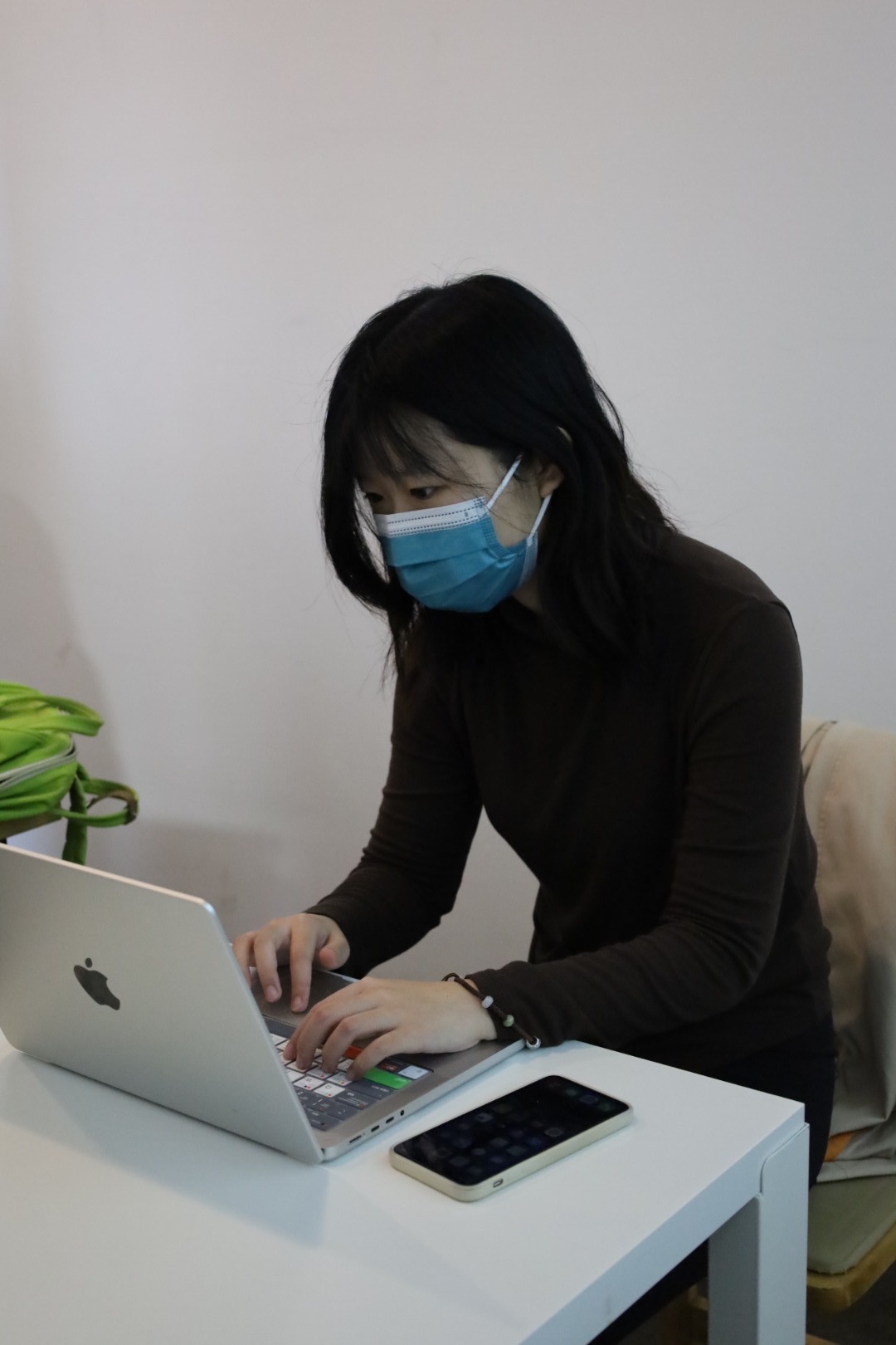 Yiwen Xu, who has shoulder-length straight hair with bangs, is wearing a surgical mask and all black outfit. She is working on her laptop on a white desk.