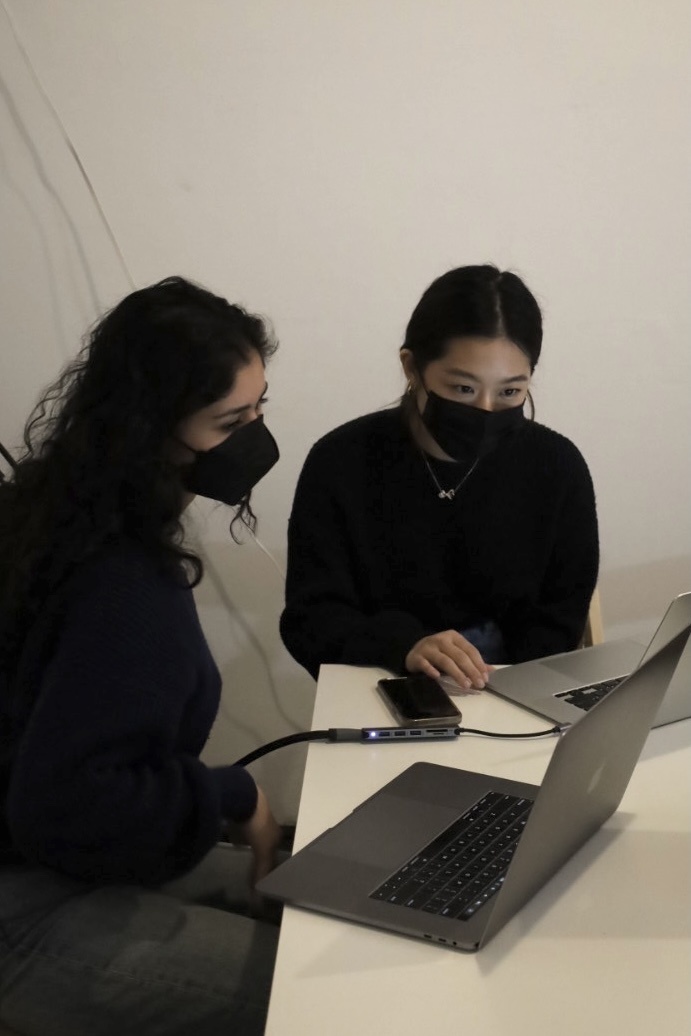 Zoyah Shah, who has shoulder-length, dark brown curly hair and Gina Lee, who has straight black hair tied to a low ponytail, are both wearing black masks and black long-sleeves shirts. They are sitting at a white desk and sharing a laptop screen.