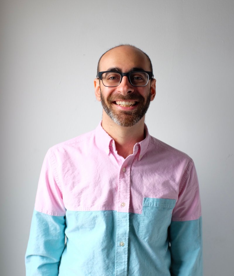 A person looking at the camera and smiling. They are wearing a pink and blue shirt and standing in front of a white screen.