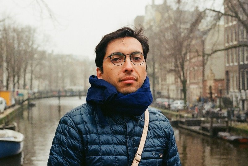 A person in a jacket and scarf looking at the camera. They seem to be standing on a bridge over a river. Tree and apartments are in the background.