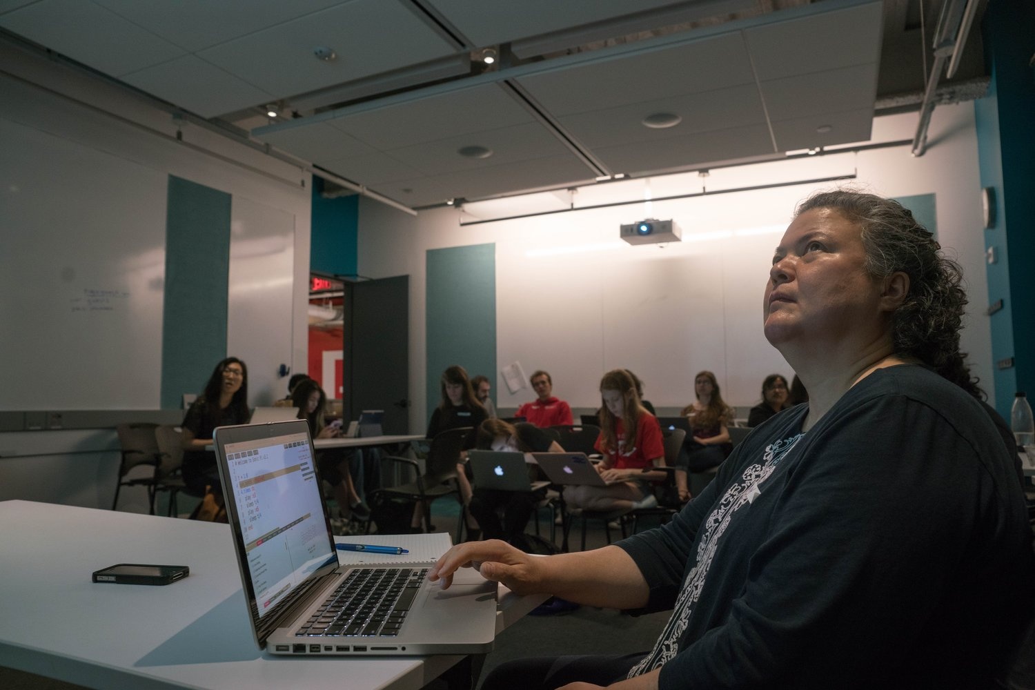 A person with a laptop looking up at the projection screen in a classroom. There a group of people sitting behind her in desks.