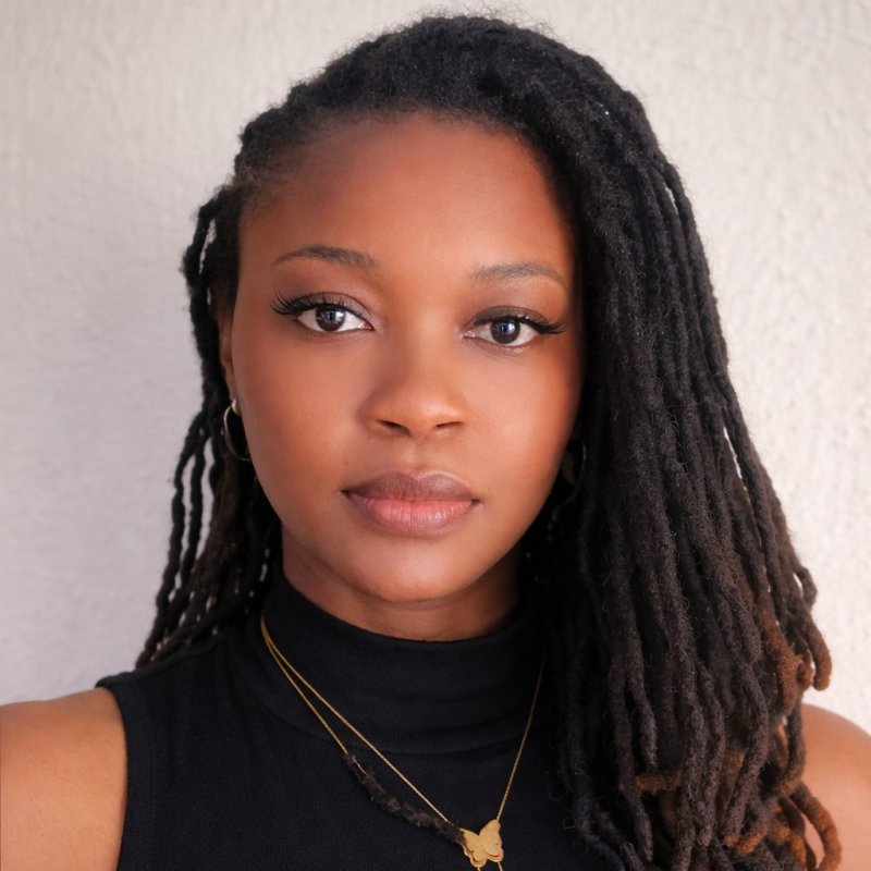 Headshot of Nwakaego Ego, a young woman with locs wearing a black sleeveless turtleneck and a gold butterfly necklace, photographed against a light grey background.