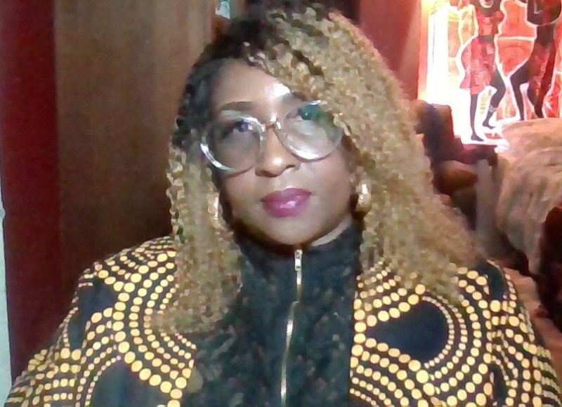 A dark-skinned African woman with black to blonde curly hair, wearing clear-rimmed glasses,  a black lace top, as well as a black jacket with gold dots. She is sitting in front of a red wall, with a mural featuring dark-skinned African dancers in red attire.