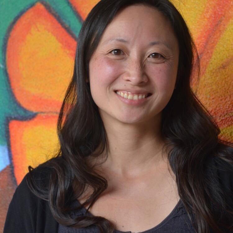 Angi, an Asian woman, is smiling at the camera. She wears a black top and has long black hair. In the background is a wall painting of a large orange flower.