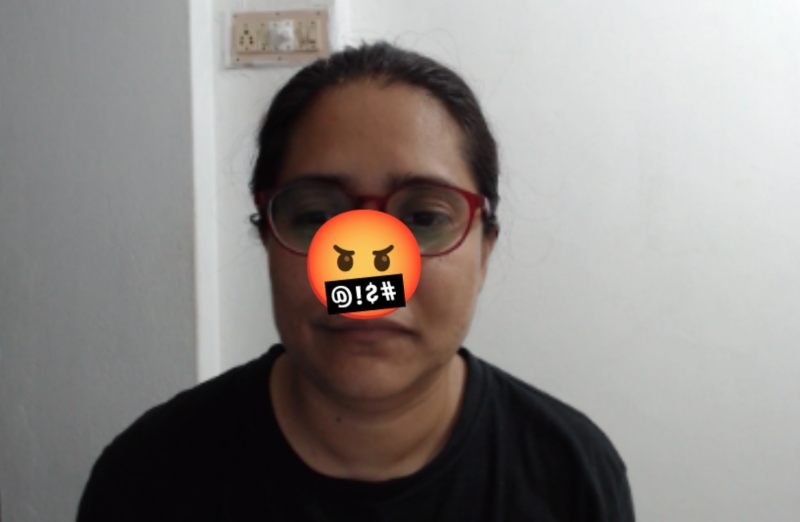 A bespectacled adult woman of Indian origin looks at the camera. The angry, cursing emoji covers part of her face.