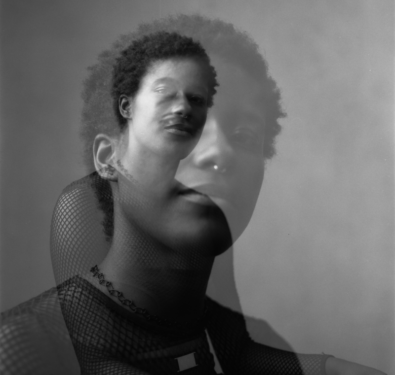 A black and white glitched double exposure of Cy, a dark-skinned black agender person with short black hair turning slightly toward the camera. They are wearing a black mesh shirt and black choker necklace.