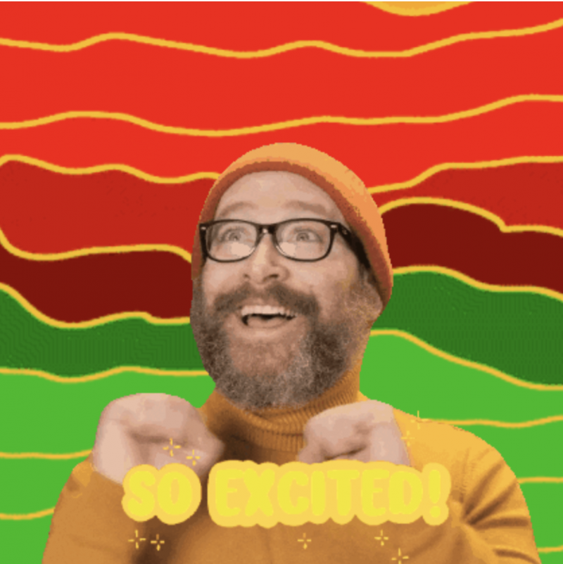 Shawn Higgins, a bearded white male with a red wool cap and black glasses shakes his hands in excitement. He wears a yellow turtleneck sweater and smiles. His beard is large and puffy, brown but flecked with grey. In front of him the words SO EXCITED flash in orange. Behind him is a rainbow of shades of red and green.