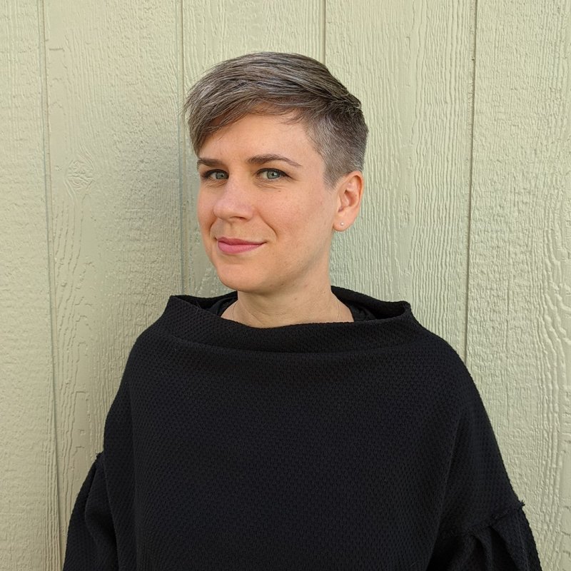 Kate is wearing a black sweater, standing in front of some light green paneling. She has pale skin and short, cropped gray hair. She looks at the camera with a shy smile.