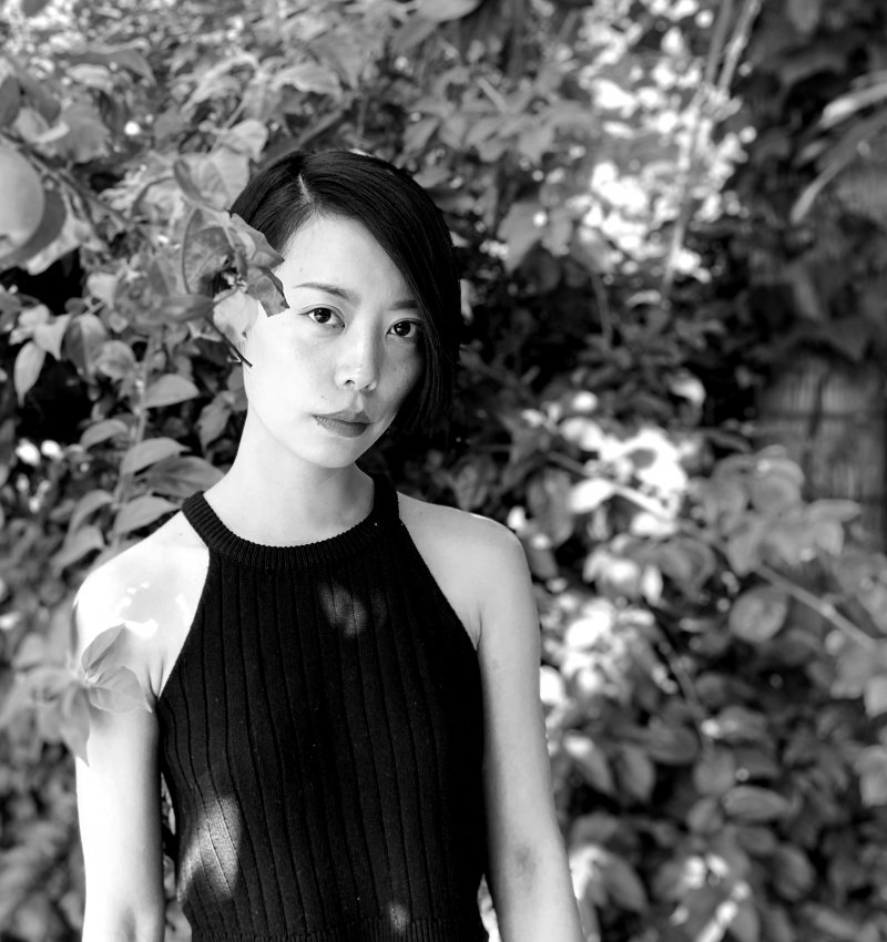 Qianqian, a non-binary Chinese person with black short hair, wearing a black tank top, standing next to an apple tree, with a lush garden in the background.