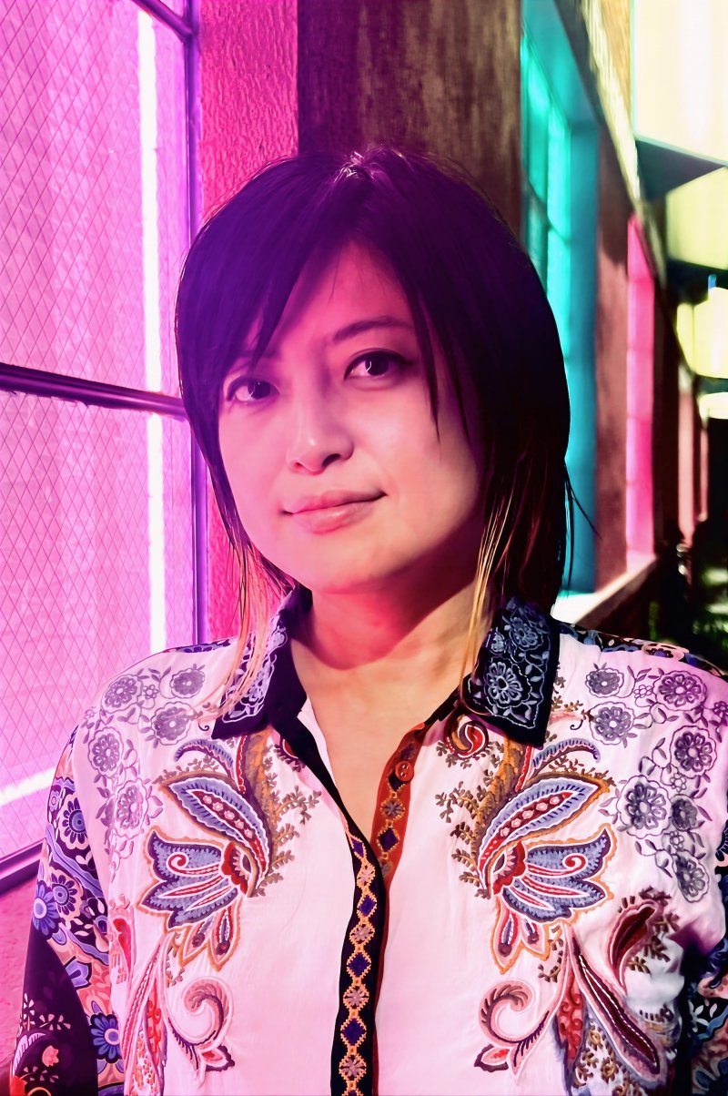 A person with black hair and blonde highlights stand in front of the Out of the Closet thrift store. They wear an ornate white button-up shirt featuring colorful paisley embroidery in blue, red, and orange tones, with a dark floral-patterned collar.